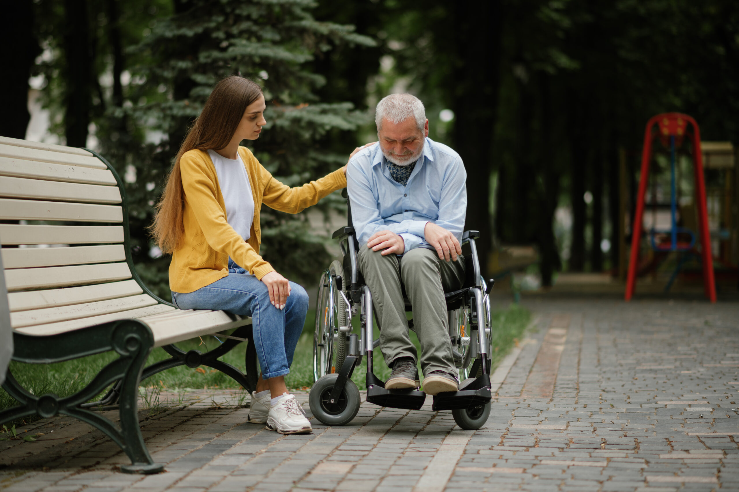 Woman takes care of a disabled father in wheelchair, family walking in park. Paralyzed people and disability, handicap overcoming. Handicapped male person and young female guardian in public place